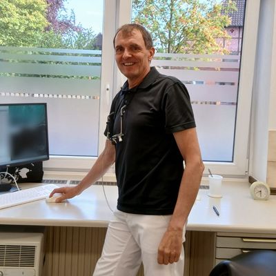 Zahnarzt Raimund Hegge steht lächelnd an seinem Arbeitsplatz vor dem Fenster
