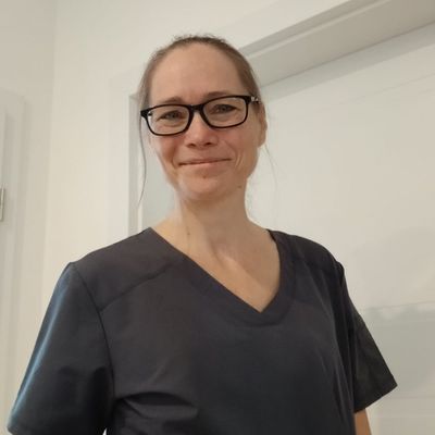 Dr. Janet Krohn mit Brille und dunkler Praxiskleidung, freundlich lächelnd